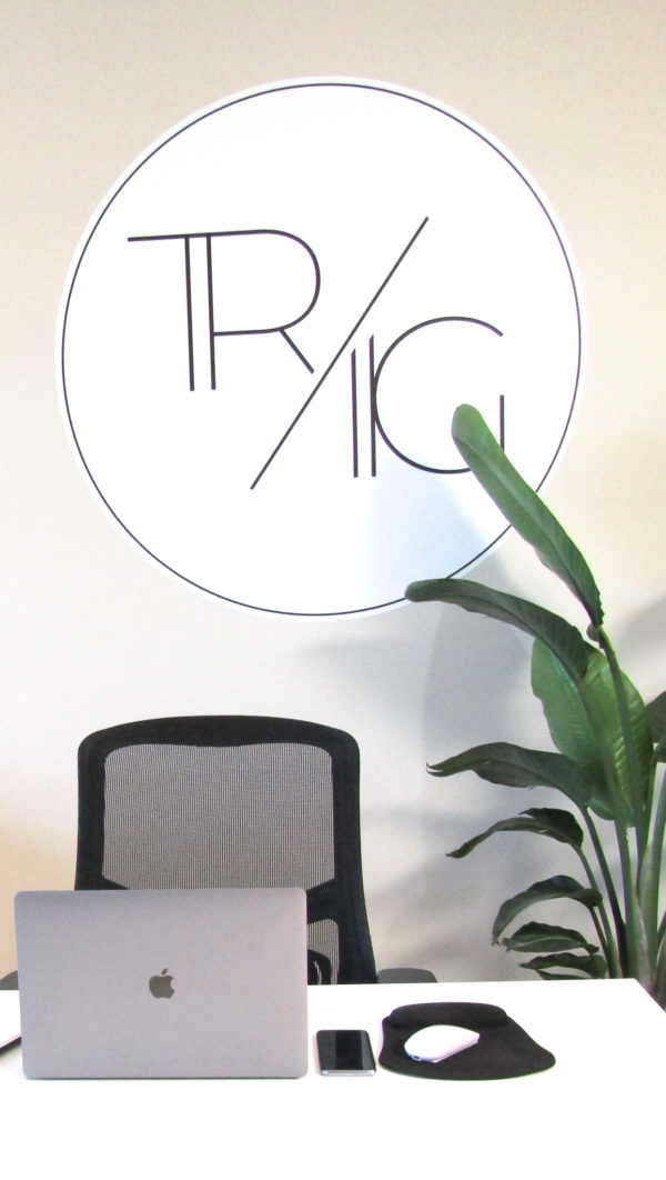 White desk with laptop and mouse pad on top and plant in the background trig logo on the wall